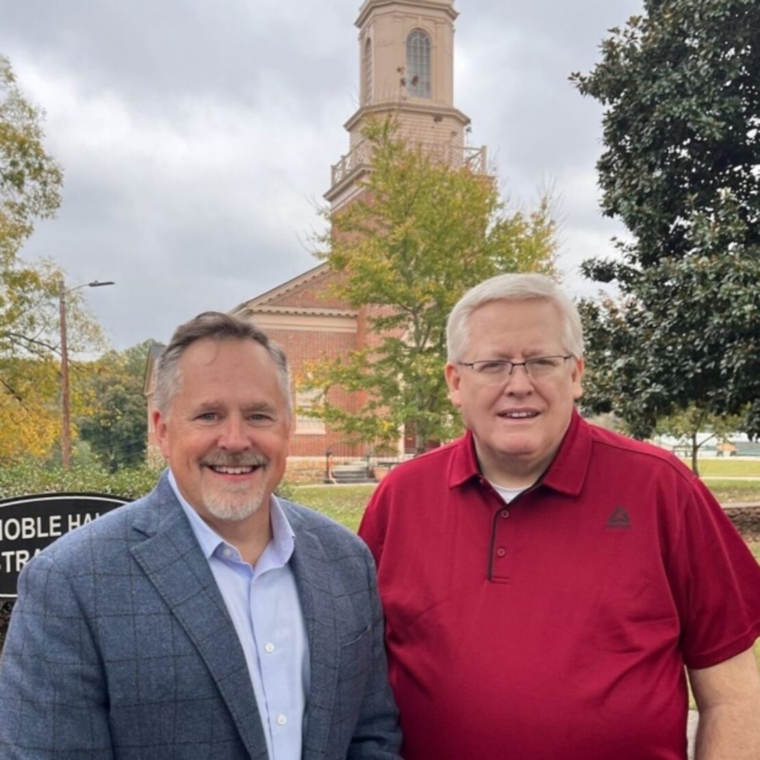 Two men stand outdoors smiling in front of a red brick church with a tall steeple. One man wears a blue blazer and the other a red polo shirt. Trees and a cloudy sky are in the background.