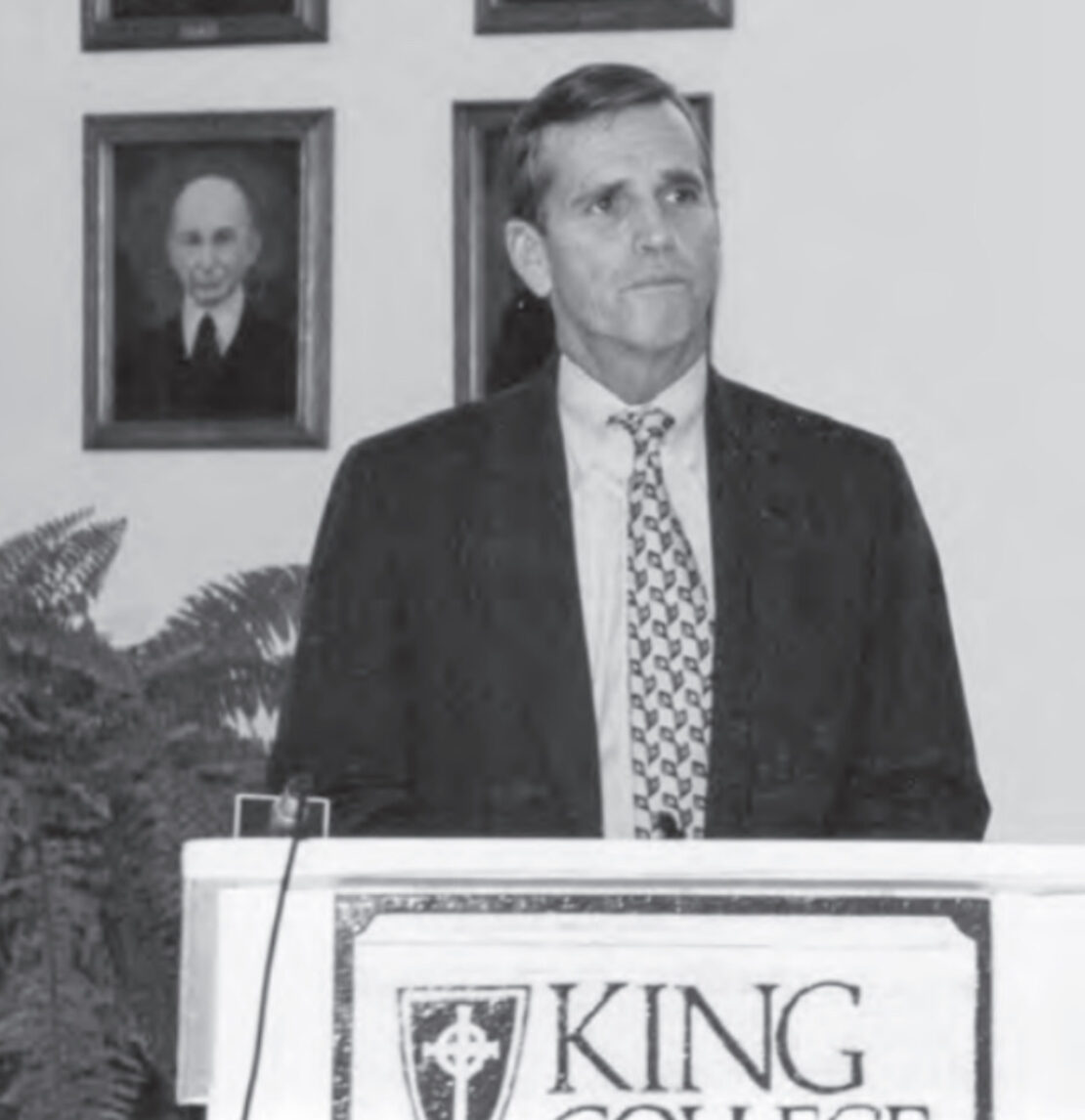 A man in a suit stands at a podium labeled King College, speaking or presenting. There are framed portraits on the wall behind him and a plant to the side.