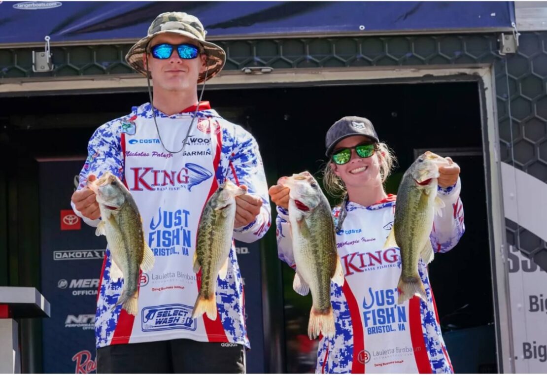 Two people wearing matching fishing jerseys and sunglasses stand side by side, each holding up two large fish, smiling at the camera in front of a fishing tournament stage backdrop.