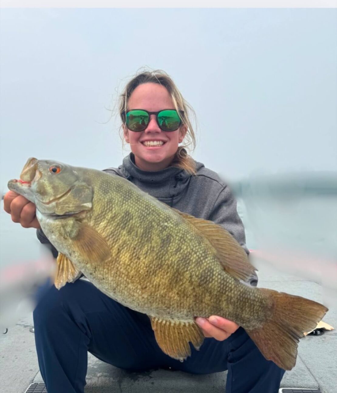A smiling person wearing sunglasses and a gray jacket holds a large fish with both hands while sitting on a boat, with a foggy body of water in the background.