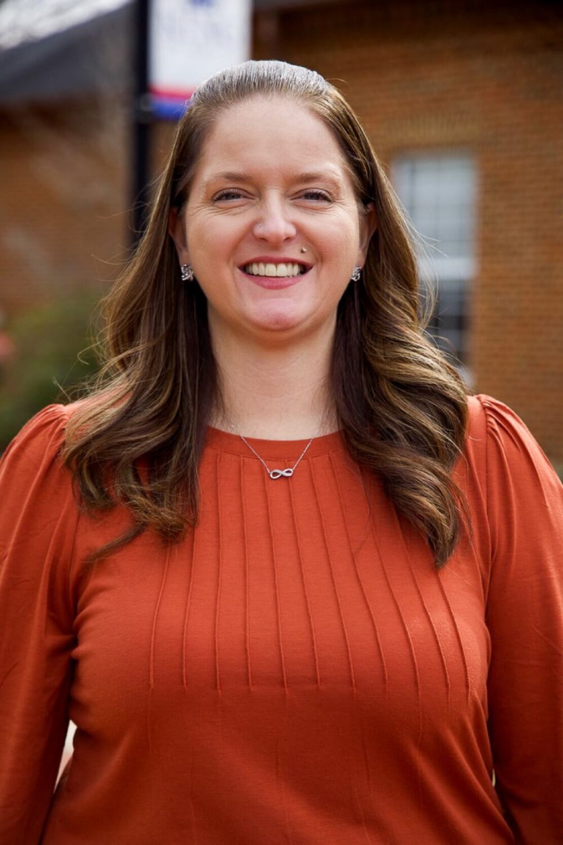 A woman with long brown hair, wearing an orange blouse and an infinity necklace, smiles while standing outside in front of a brick building.