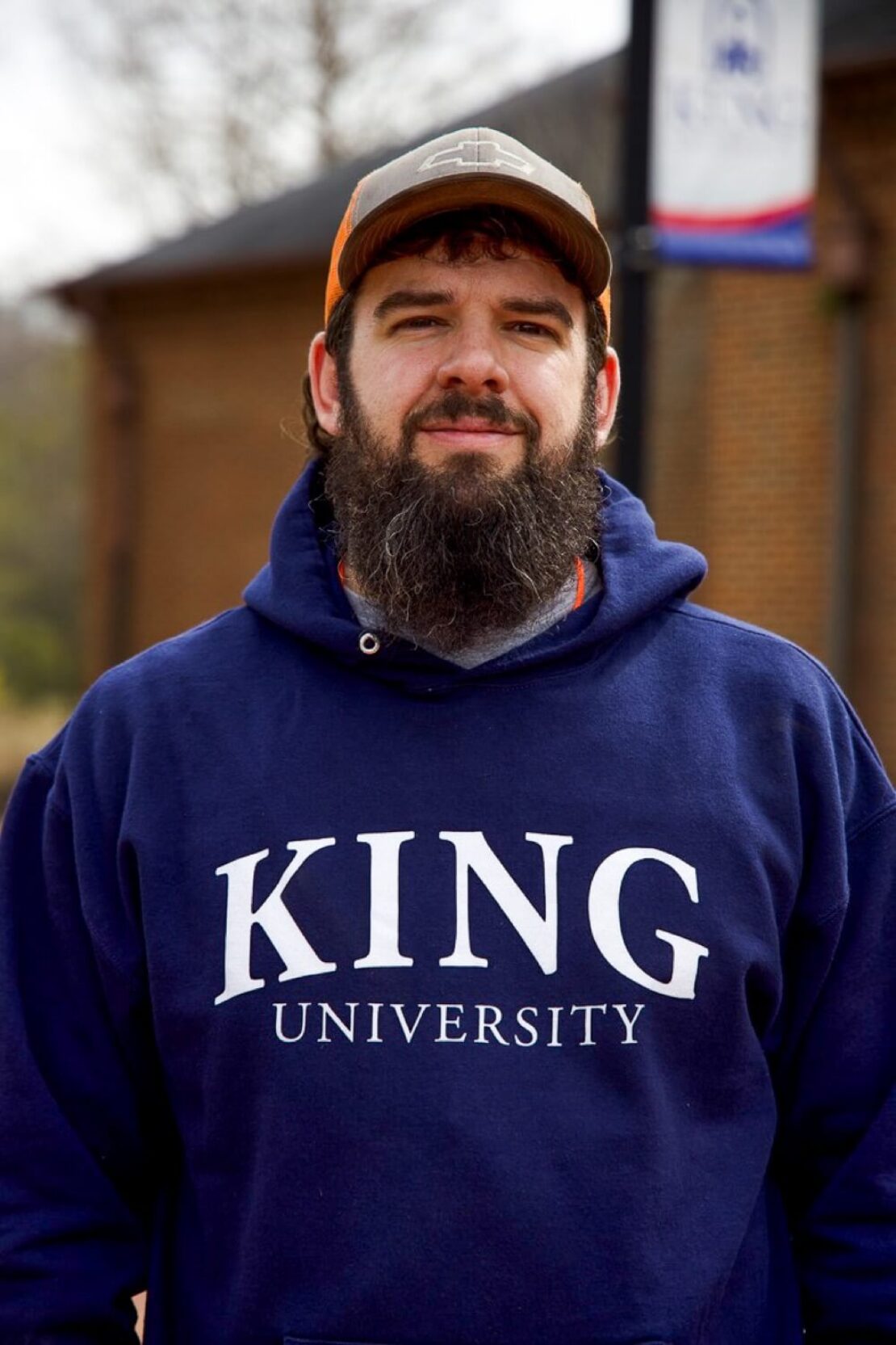 A bearded man wearing a navy blue King University hoodie and a cap stands outside in front of a brick building and a university banner.
