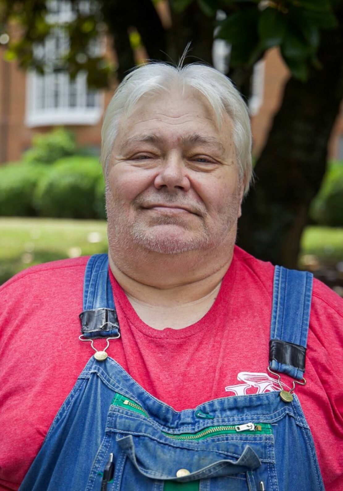 An older man with white hair and a short beard wears a red T-shirt and denim overalls, standing outdoors with greenery and trees in the background. He looks at the camera with a slight smile.