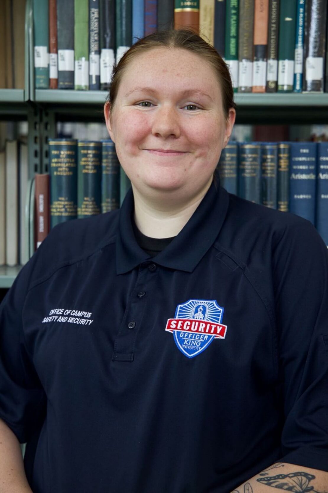 A person wearing a dark security uniform with a badge that reads Security stands in front of bookshelves filled with books, smiling at the camera.
